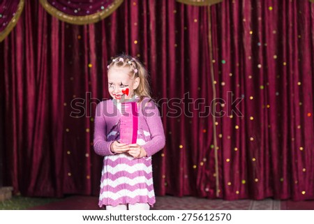 Smiling Blond Girl Wearing Clown Make Up and Striped Dress Standing on Stage Holding Over Sized Pink Comb in front of Red Curtain