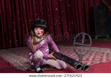 Girl Wearing Combat Helmet Crouching on Ground with Large Double Barreled Shot Gun with Iron Sight on Stage with Red Curtain
