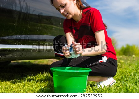 Close Up of Woman Wearing Red T-Shirt Washing Black Vehicle in Field with Sponge, Crouching Next to Green Bucket Filled with Soapy Water