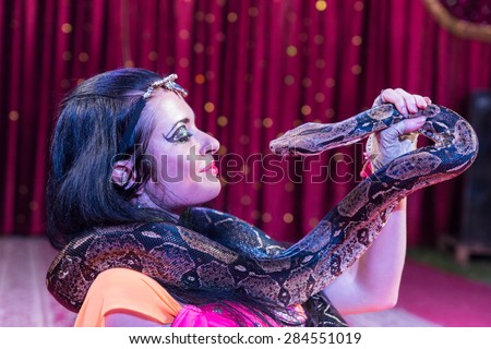 Close Up Profile of Exotic Dark Haired Snake Charmer Face to Face with Large Snake Lying on Stage with Red Curtain in Background