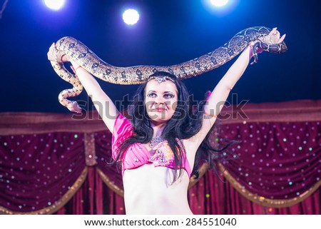 Waist Up of Exotic Dark Haired Belly Dancer Standing with Arms Raised Holding Large Snake Over Head Standing on Stage with Red Curtain in Background