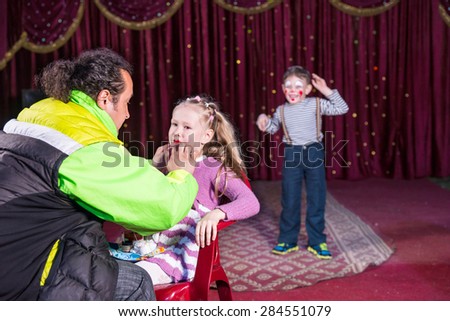 Man Applying Make Up to Face of Blond Girl Sitting in Chair with Young Boy Clown Performing in Background on Stage with Red Curtain