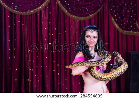 Exotic Dark Haired Belly Dancer Holding Large Snake on Stage with Red Curtain in Background
