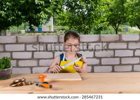 Serious Young Girl Wearing Apron, Sitting at Wooden Table with Canning Supplies and Testing Sharpness of Knife