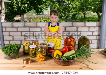 Portrait of Stern Looking Girl Standing with Arms Crossed by Wooden Table Covered with Fresh Vegetables and Jars of Pickled Preserves Outdoors by Brick Garden Wall