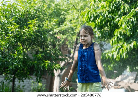 Young Girl Wearing Summer Clothes Cooling Off in Sprinkler on Warm Summer Day, Looking Surprised by Spray of Water on Back