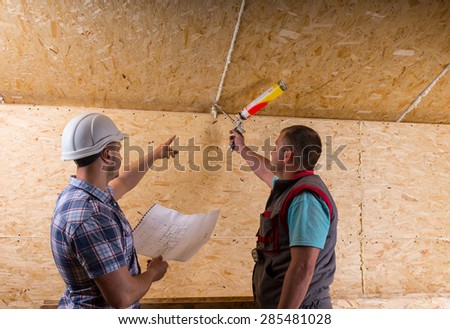 Foreman Wearing White Hard Hat and Holding Building Plans Instructing Worker How to Apply Caulk to Seam in Unfinished Wood Ceiling in New Home