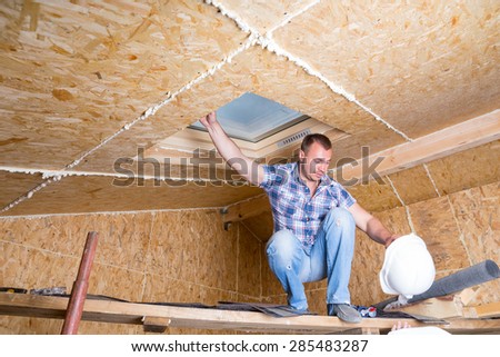 Male Construction Worker Builder Holding White Hard Hat and Balancing on Wooden Plank Scaffolding near Ceiling of Unfinished Home with Exposed Particle Plywood Board