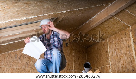 Male Construction Worker Builder Wearing White Hard Hat Crouching on Elevated Scaffolding near Ceiling Sky Light and Reading Plans in Unfinished Home with Exposed Plywood Particle Board