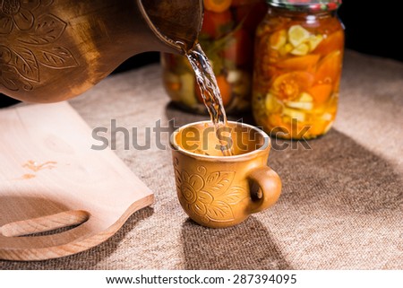 Close Up of Water Pouring into Carved Wooden Drinking Cup from Pitcher on Burlap Covered Table with Jars of Pickled Vegetable Preserves and Wooden Cutting Board in Background