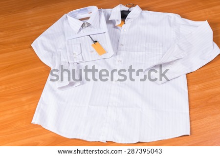 Mens White Dress Shirt Laid Out Flat on Wooden Surface, with Identical Shirt Folded with Tag Attached on Wooden Background