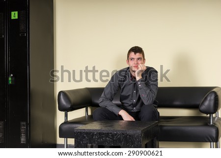 Bored or anxious man sitting on a modern black leather settee in a waiting room with is chin on his hand staring morosely to the side