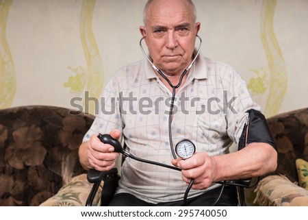 Senior Bald Man with Special Needs, Sitting on his Wheelchair and Holding Sphygmomanometer and Stethoscope Apparatus, Looking Straight at the Camera.