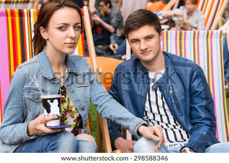 Young Woman with Mobile and Coffee Sitting in Striped Beach Chair with Hand on Knee of Young Man at Outdoor Picnic or Festival
