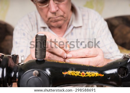 Elderly man wearing glasses threading a needle with yarn as he prepares to do his sewing on his sewing machine