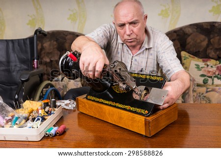 Senior Man Inspecting Bottom of Old Fashioned Sewing Machine in Living Room at Home with Variety of Threads and Supplies on Table