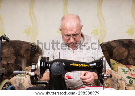 Serious Senior Man Looking Down at Spool of Red Thread and Threading Old Fashioned Manual Sewing Machine at Home in Living Room