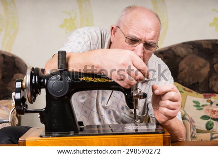 Senior man wearing glasses sitting in his living room using a sewing machine carefully threading the needle with thread