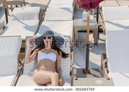 Stylish young woman lying sun tanning lying in her bikini, straw sunhat and trendy sunglasses on a recliner chair at a seaside resort
