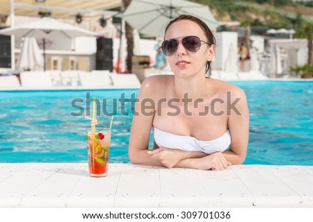 Sexy Young Woman with Sunglasses Smiling at the Camera While Leaning on the Edge of Swimming Pool with a Glass of Juice on the Side.