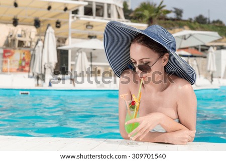 Elegant young woman standing in the water of a resort swimming pool sipping an ice cold cocktail from a glass on the tiled surround on her summer vacation