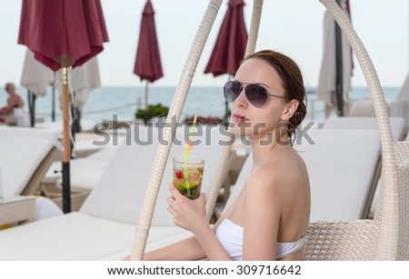Young Woman in White Bikini Holding Tropical Drink and Looking Back Over Shoulder While Sitting in Wicker Chair on Oceanfront Deck at Luxury Vacation Resort