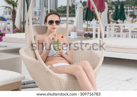 Young Woman Wearing White Bikini and Dark Sunglasses Relaxing in Wicker Deck Chair with Tropical Drink on Patio of Oceanfront Luxury Vacation Resort
