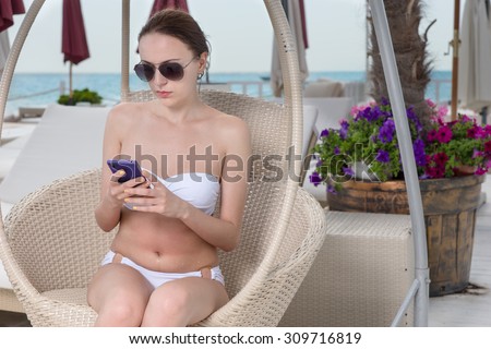 Young Woman Wearing White Bikini and Dark Sunglasses Sitting in Hanging Wicker Chair on Pool Deck of Luxury Resort Looking Down at Cell Phone and Texting