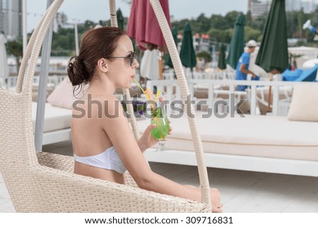 Young Woman in White Bikini Sipping Tropical Drink Through Straw While Sitting in Wicker Chair on Oceanfront Deck at Luxury Vacation Resort