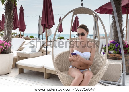 Young Woman Wearing White Bikini and Dark Sunglasses Sitting in Hanging Wicker Chair on Pool Deck of Luxury Resort Looking Down at Cell Phone and Texting