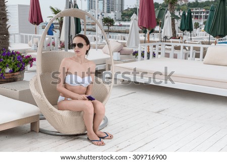 Young Woman Wearing White Bikini and Dark Sunglasses Holding Cell Phone and Sitting Anxiously in Hanging Wicker Chair on Pool Deck of Luxury Resort