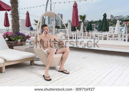 Full Length of Young Man in Bathing Suit Making Call on Cell Phone While Relaxing in Wicker Deck Chair on Patio of Oceanfront Luxury Vacation Resort