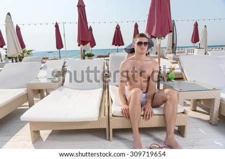 Young Man Wearing Sunglasses Sitting on Edge of Lounge Chair on Deck of Oceanfront Luxury Beach Resort