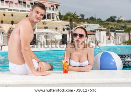Portrait of Young Couple with Tropical Drink Relaxing on Edge of Outdoor Swimming Pool at Luxury Tropical Resort