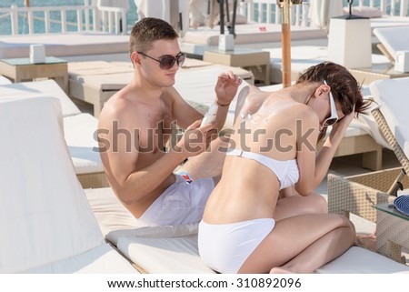 Handsome Boyfriend Putting Sunscreen Lotion on the Back of his Girlfriend While Sitting on the Lounge Chair in the Resort, on a Tropical Climate.
