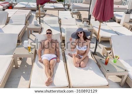High Angle View of Young Couple with Tropical Drinks Relaxing on Lounge Chairs on Sunny Deck of Oceanfront Luxury Beach Resort