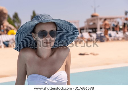 Waist Up Close Up of Young Woman on Vacation Wearing Sun Hat, Sunglasses and White Bikini While Sitting on Sunny Deck of Public Resort Swimming Pool