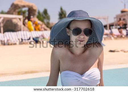 Waist Up Close Up of Young Woman on Vacation Wearing Sun Hat, Sunglasses and White Bikini While Sitting on Sunny Deck of Public Resort Swimming Pool