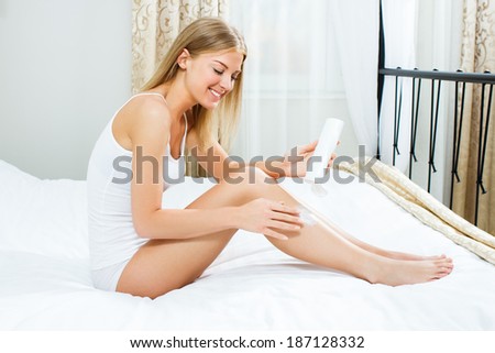 Blonde woman is  sitting on bed in her bedroom  and putting lotion on her body,Skin care