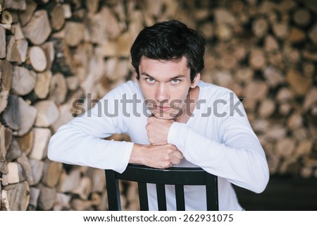 Young man sitting and leaning on the back of the chair. Serious look. Firewood background.
