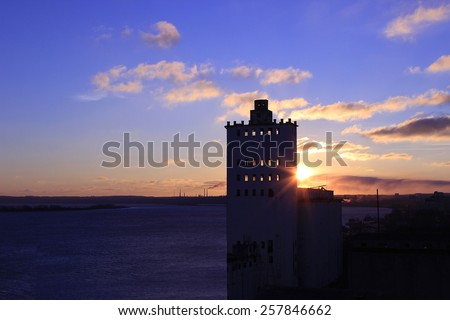 urban grain elevator in the rising sun near the factory buildings of the city in a snow-covered ice of the river.