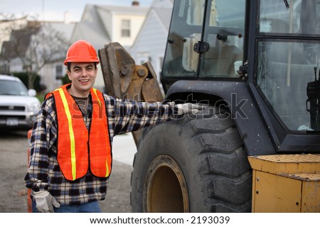 Construction road worker, standing next to tractor/bulldozer/excavator, his hand on the wheel. Residential area on the background.