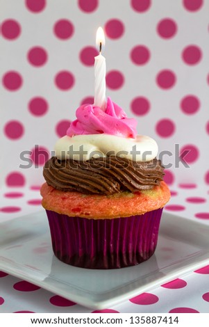 1 neapolitan frosted cupcake on white plate with pink polka dot background and 1 lit birthday candle