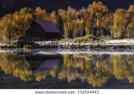 Fall scene of an abandoned barn reflected in lake with changing fall trees and frost on ground