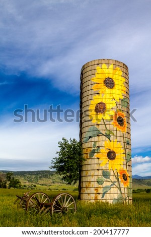 Rural farm silo with painted sunflowers and old farm equipment on sunny summer morning in mountain foothills