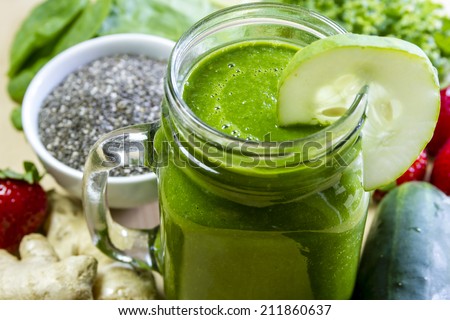 Healthy green juice smoothie surrounded by whole fruits and vegetables with cucumber garnish