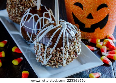 Hand dipped caramel apples with nuts and chocolate drizzle sitting on white plate with halloween pumpkin bucket and candy corn