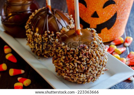 Homemade caramel apples with nuts and chocolate drizzle sitting on white plate with halloween pumpkin bucket and candy corn