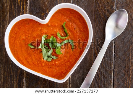 Homemade tomato and basil soup in white heart shaped bowl with spoon on wooden table