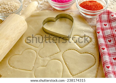 Heart shaped cookie cutters cutting out holiday sugar cookies with wooden rolling pin and red and pink sugar sprinkles sitting next to red heart napkin